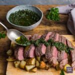 Slices of medium-rare steak positioned over a pile of cubed rosemary potatoes with a herb and chilli dressing being spooned over the top.