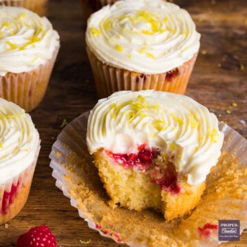 Raspberry muffin topped with lemon butter cream sitting on a muffin case with the sides pulled away and surrounded a few other muffins.