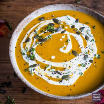 Bowl of pumpkin soup topped with a swirl of cream and a sprinkle of pumpkin seeds and parsley.