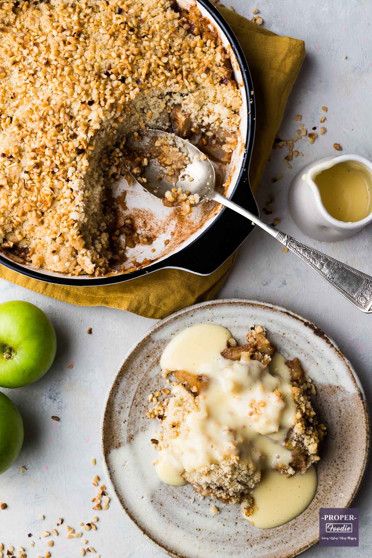 A portion of apply crumble on a small plate with custard drizzled over and the rest of the crumble in a larger dish with a spoon.