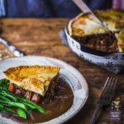 A slice of steak and ale pie on a plate with gravy and tender-stem broccoli with pie dish in the background.