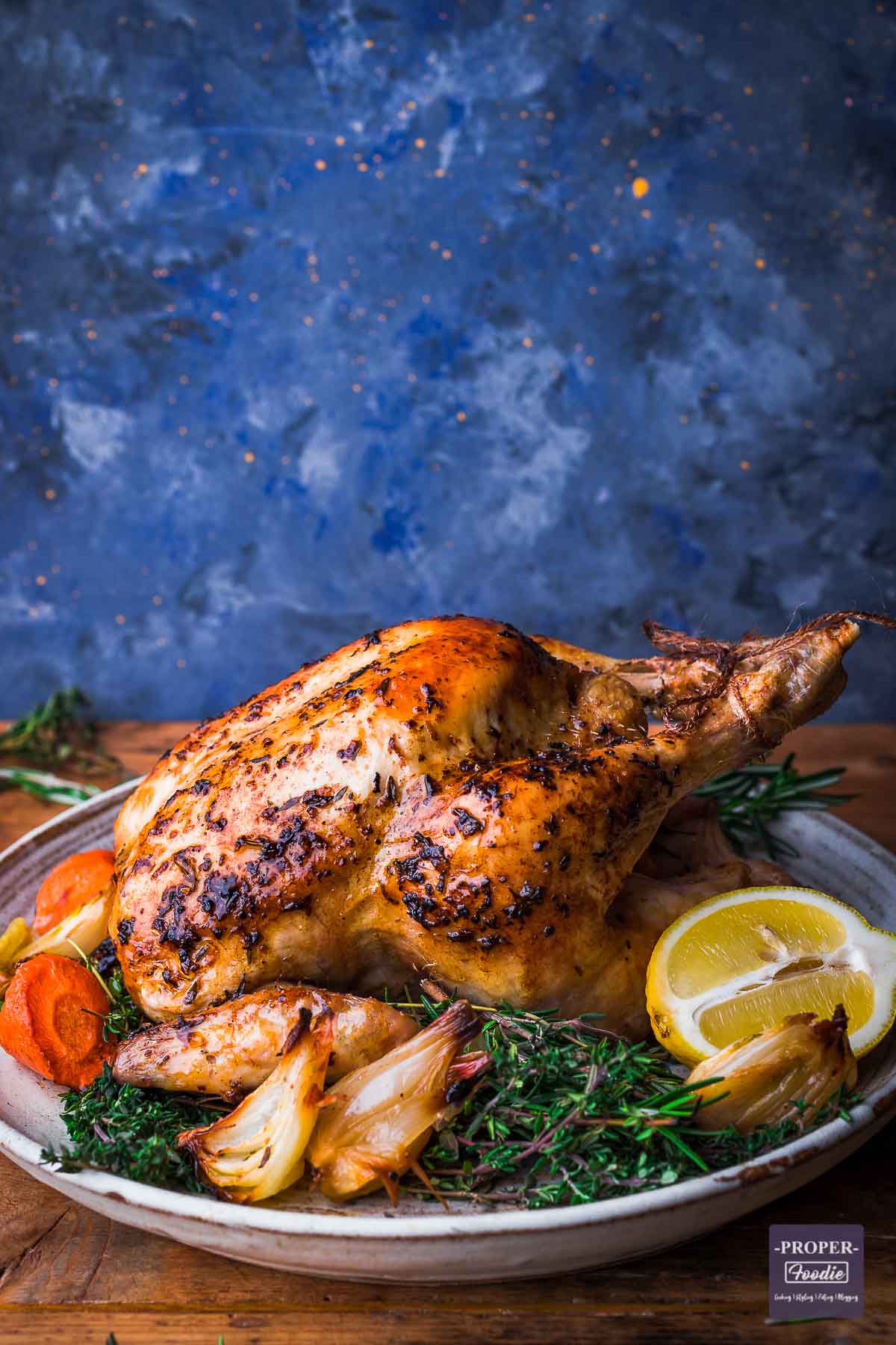 Whole roast chicken with golden skin served on a large plate surrounded by roast carrots, onions and fresh herbs.