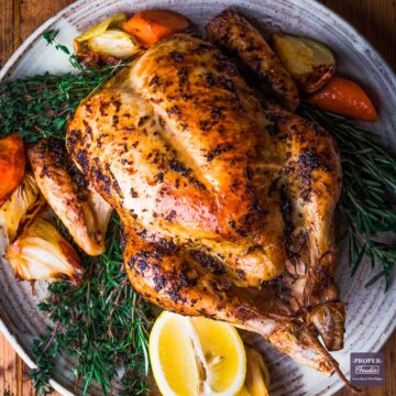 whole chicken roasted and presented on a large plate with cooked carrots and onions and fresh herbs.