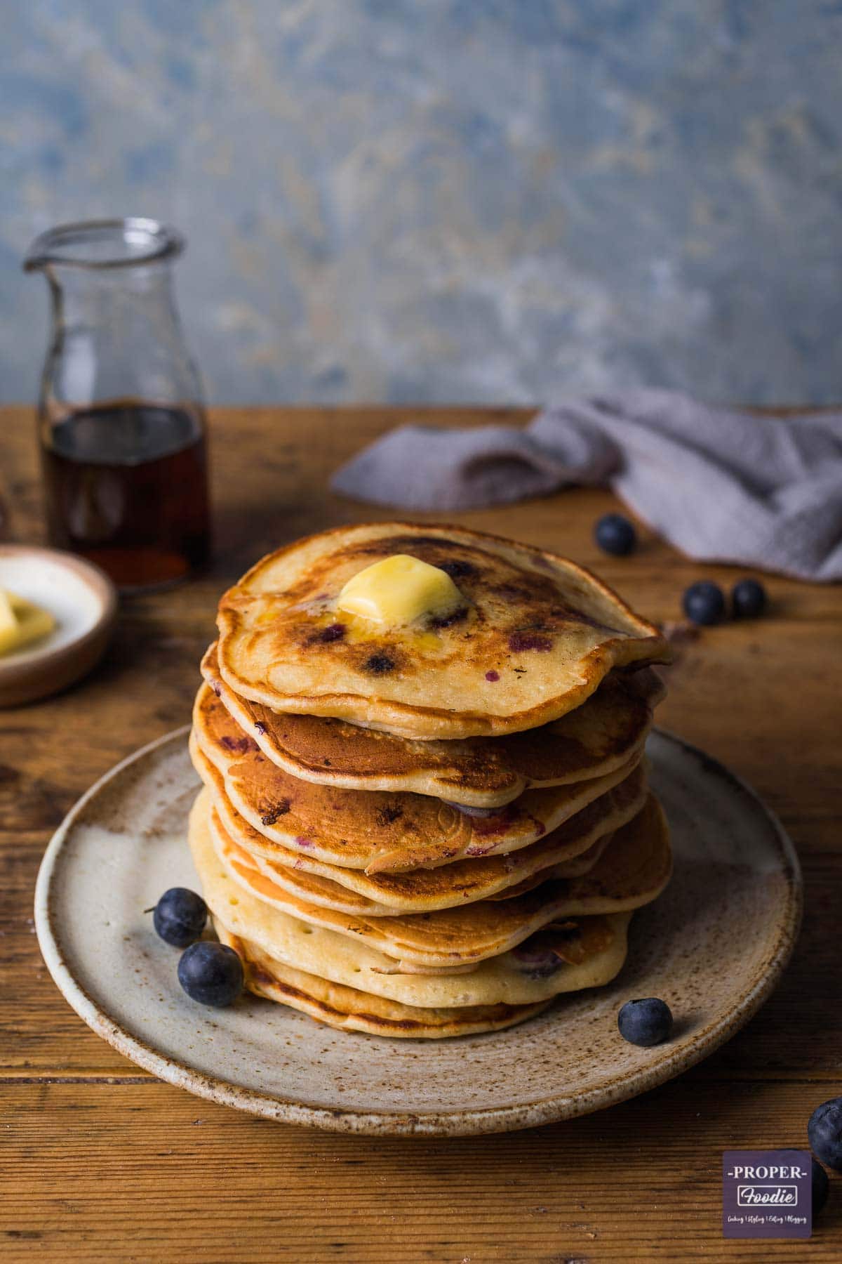 Stack of blueberry pancakes with butter melting on the top and a small glass jug of syrup in the background