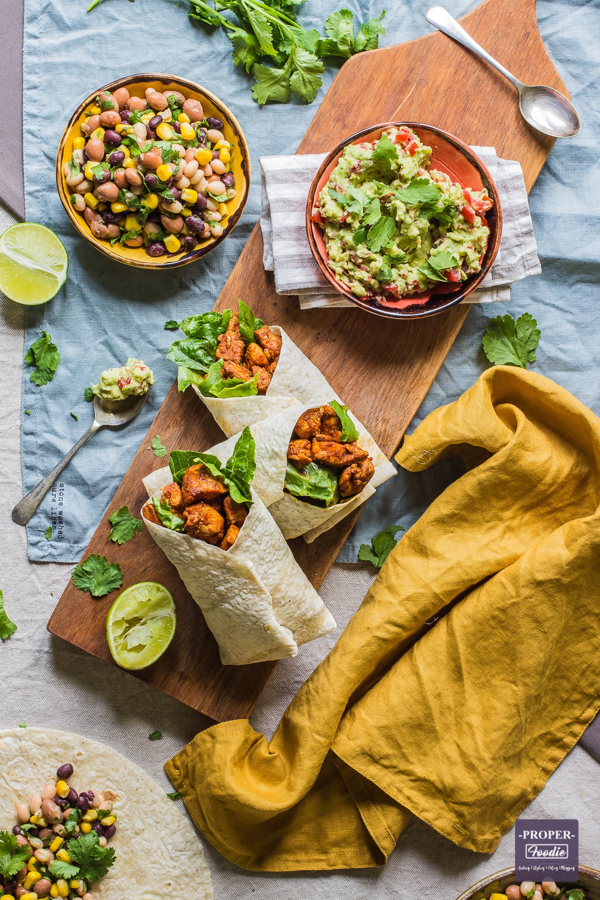 three chicken wraps on a board viewed from above with half a lime squeezed at the side and small bowls of bean salad and smashed avocado above