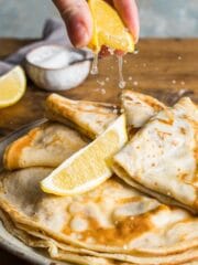 A plate of this flat pancakes stacked with the top four folded twice topped with a lemon wedge and a second wedge being squeezed over the top.