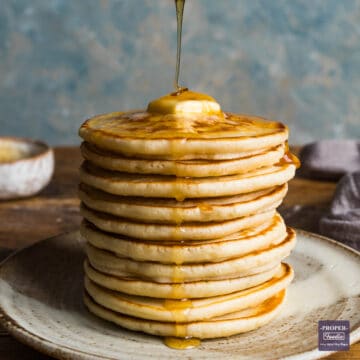A tall stack of scotch pancakes on a plate with melted butter on top and honey being drizzled over with a wooden honey dipper.